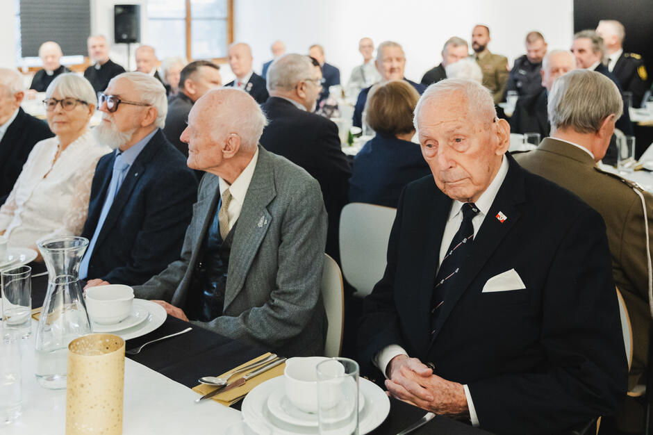 Na zdjęciu widać elegancko ubranych starszych ludzi siedzących przy odświętnie przygotowanym stole podczas formalnego spotkania. Atmosfera sugeruje uroczysty charakter wydarzenia, z udziałem osób o poważnym i refleksyjnym wyrazie twarzy, być może związanym z uhonorowaniem lub upamiętnieniem.