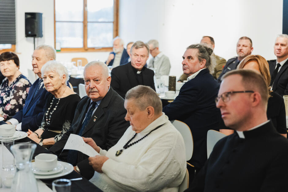 Na zdjęciu widać grupę elegancko ubranych osób siedzących przy odświętnie nakrytym stole, co sugeruje formalne spotkanie lub uroczystość. Wśród uczestników znajdują się zarówno osoby duchowne, jak i cywilne, a jedna z kobiet czyta dokument, co może wskazywać na oficjalny charakter wydarzenia.