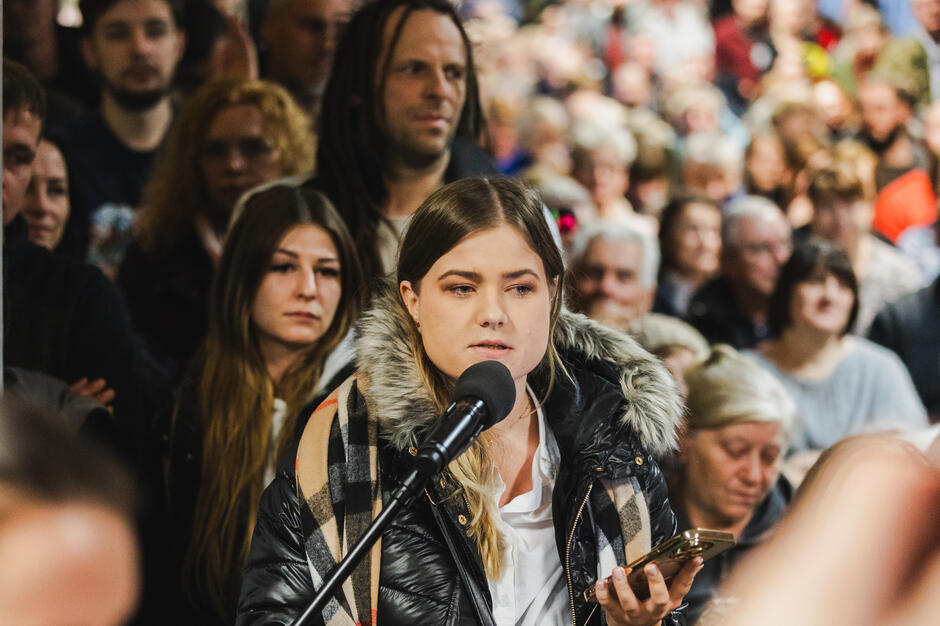 na zdjęciu młoda kobieta w kurtce, mówi do mikrofonu, wokół niej tłum ludzi