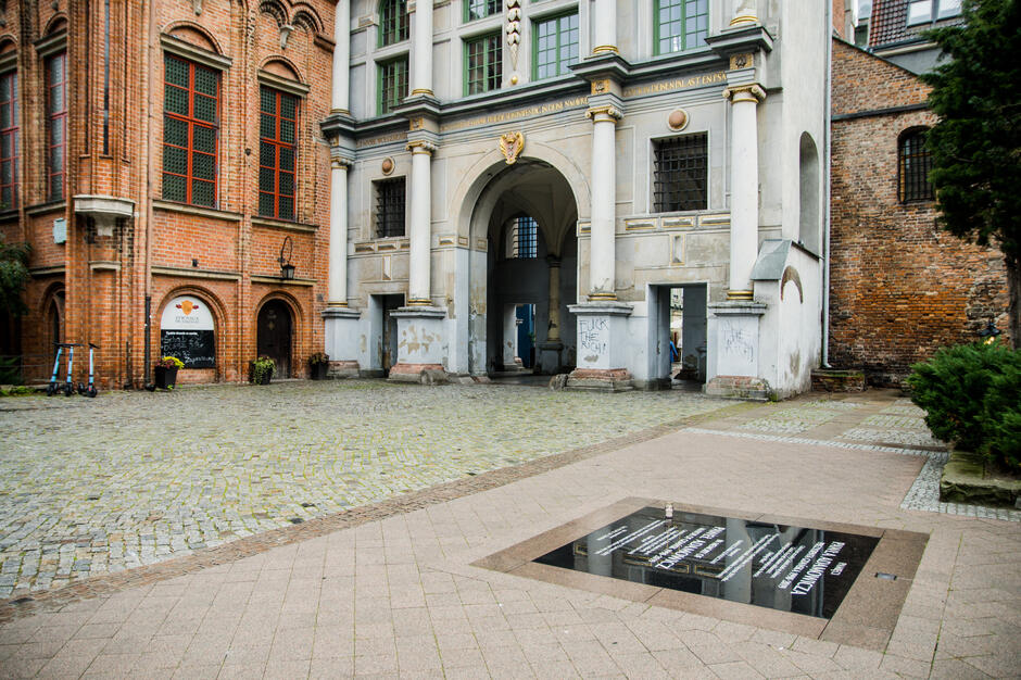 Zdjęcie przedstawia fragment historycznego budynku w Gdańsku, w którego otoczeniu widoczna jest tablica pamiątkowa poświęcona Pawłowi Adamowiczowi. Tablica znajduje się na tle zabytkowych murów i łuku, podkreślających charakter miejsca, w którym oddano hołd zmarłemu prezydentowi miasta