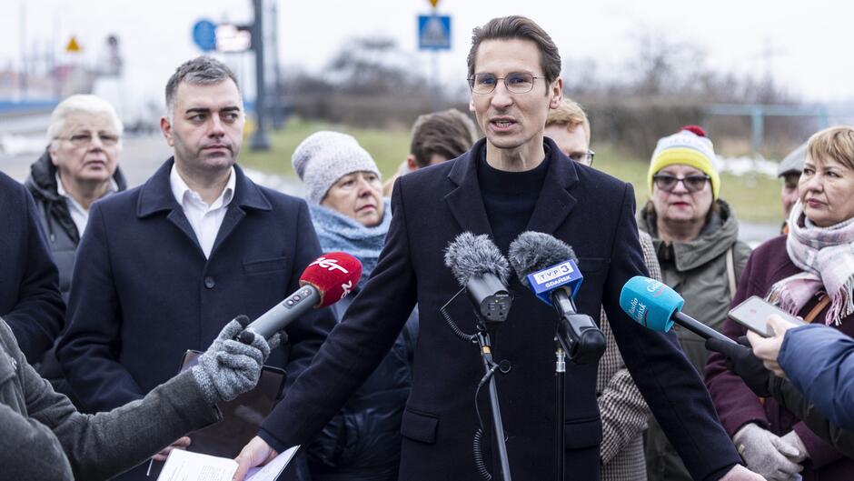 Na zdjęciu widać konferencję prasową na świeżym powietrzu, w której uczestniczy kilku dorosłych ludzi. Na pierwszym planie znajduje się mężczyzna w okularach, ubraną w ciemny płaszcz, przemawiający do mikrofonów różnych mediów. Przed nim widać wyciągnięte ręce dziennikarzy trzymających mikrofony i dokumenty. W tle stoi grupa osób, prawdopodobnie uczestników spotkania, ubranych w ciepłe zimowe ubrania, w tym czapki i szaliki. Scena odbywa się w chłodny, pochmurny dzień na zewnątrz, w okolicy drogi i zielonego terenu. Widać również oznaczenia drogowe.