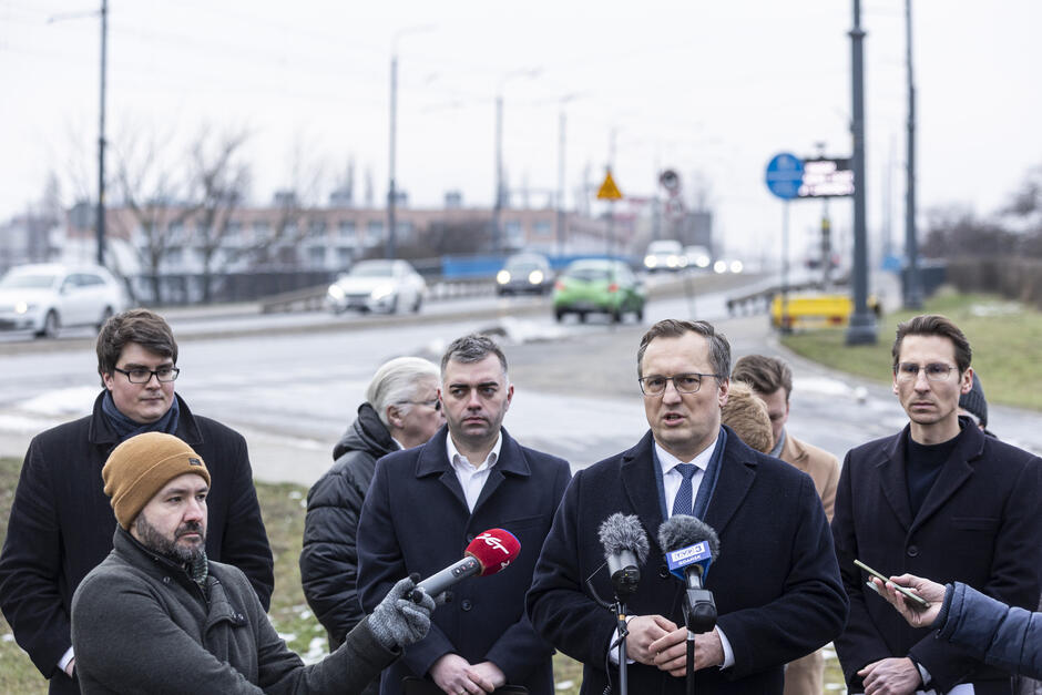 Na zdjęciu przedstawiono konferencję prasową na zewnątrz, odbywającą się w zimowej scenerii. Na pierwszym planie stoi mężczyzna w okularach i garniturze, przemawiający do mikrofonów, które należą do różnych mediów. Obok niego, po prawej stronie, znajduje się inny mężczyzna w ciemnym płaszczu, a w tle widać kilka osób – część z nich ma na sobie ciepłe zimowe ubrania, takie jak czapki i szaliki. W tle widoczna jest droga z przejeżdżającymi samochodami, chodnik oraz budynki przemysłowe lub mieszkalne. Pogoda jest pochmurna, a na trawie leży resztka śniegu, co sugeruje zimową porę. Fotograf uchwycił moment rozmowy lub wystąpienia dotyczącego jakiejś lokalnej sprawy.