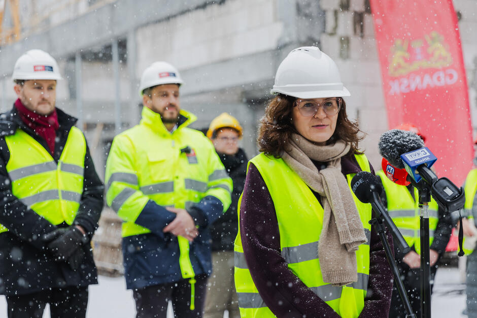 Na zdjęciu widzimy konferencję prasową na tle budowy w zimowej scenerii, podczas której przemawia kobieta w żółtej kamizelce odblaskowej i białym kasku ochronnym, stojąc przed mikrofonami z logotypami lokalnych mediów. W tle stoją inni uczestnicy wydarzenia ubrani w odzież roboczą i kaski, a także widoczne są elementy konstrukcji budynku oraz baner z napisem „Gdańsk”