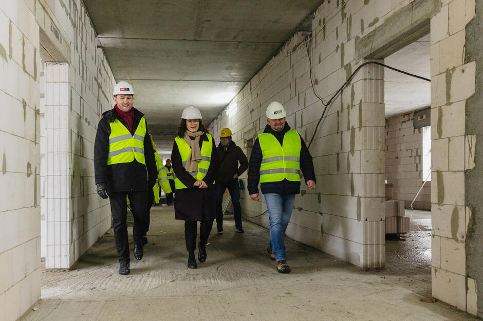 Na zdjęciu grupa osób w kaskach ochronnych i kamizelkach odblaskowych zwiedza wnętrze powstającego budynku w stanie surowym. Widoczne są surowe ściany z bloczków betonowych, a uczestnicy spacerują korytarzem, obserwując postępy budowy