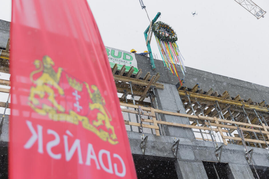 Na zdjęciu przedstawiono moment zawieszenia tradycyjnej wiechy na szczycie konstrukcji budynku w stanie surowym, co symbolizuje zakończenie tego etapu budowy. Na pierwszym planie widoczny jest czerwony baner z herbem i napisem „Gdańsk”, a w tle widoczni są pracownicy budowlani oraz wiecha ozdobiona wstążkami