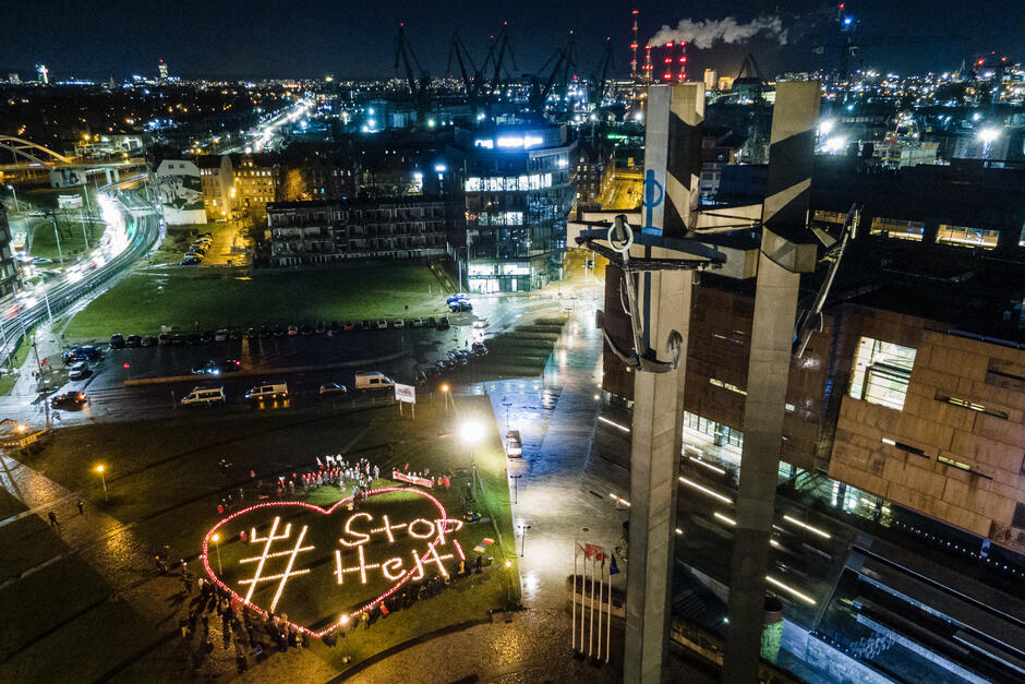 Zdjęcie z lotu ptaka. Na placu w mieście płoną znicze ułożone w kształt wielkiego serca, w środku którego ze zniczy ułożony jest napis hasztag stop hejt. Obok stoi pomnik Trzech Krzyży, w tle widać miasto i światła zapalone w budynkach. Jest wieczór.