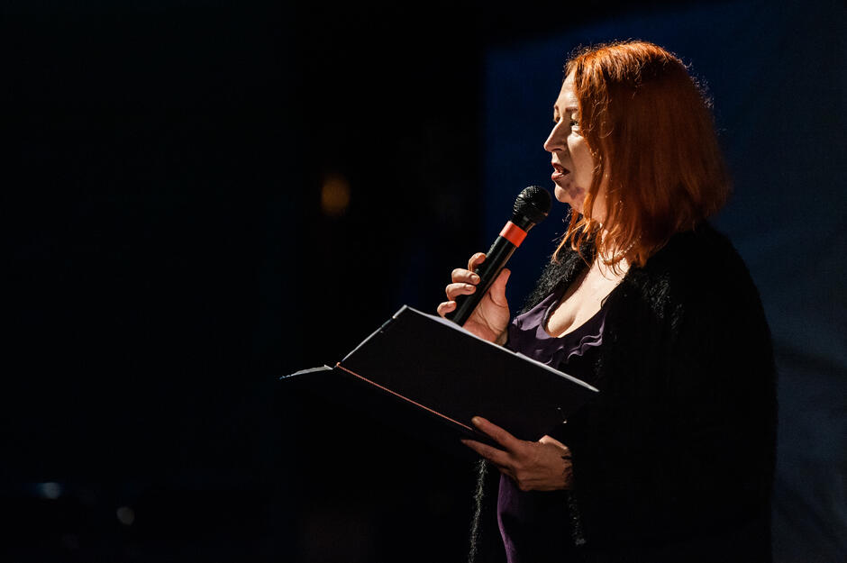 kobieta o rudych włosach czyta do mikrofonu, w rękach ma teczkę a w niej białe kartki