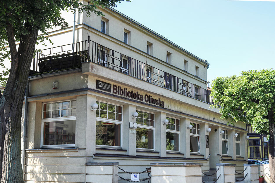 Na zdjęciu znajduje się budynek Biblioteki Oliwskiej, zlokalizowany w otoczeniu drzew i utrzymany w stylu modernistycznym. Jasne elewacje z dużymi oknami oraz balkon na piętrze nadają mu elegancki i przyjazny charakter, a widoczny szyld informuje o funkcji budynku
