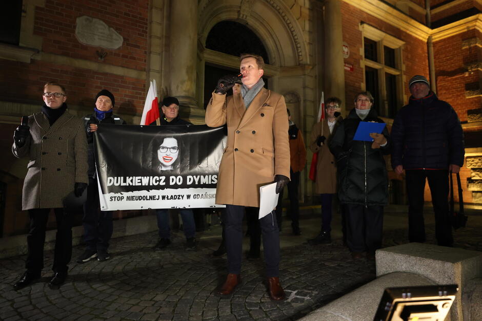 na zdjęciu manifestujące osoby, widać czarny baner z twarzą kobiety, obok stoi mężczyzna w jasnym płaszczu, przemawia do mikrofonu