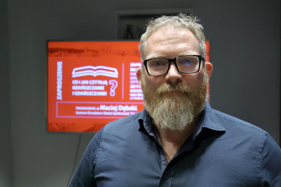 mężczyzna w ciemnej koszuli i z jasną brodą i w okularach stoi na tle czerwonej skrzynki wiszącej na ścianie