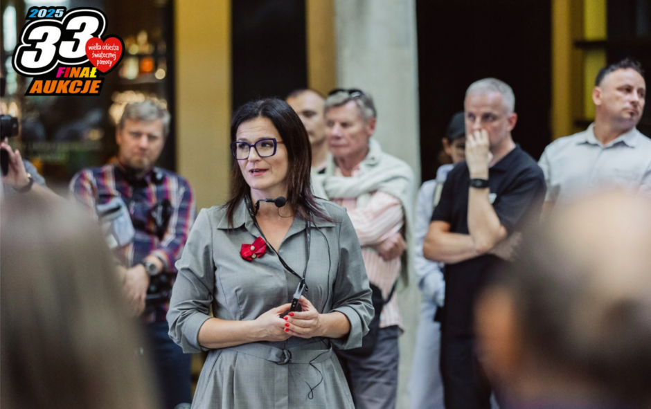 Na zdjęciu widzimy kobietę w okularach, ubraną w szarą sukienkę z przypiętym czerwonym symbolem, przemawiającą do zgromadzonych ludzi. W tle znajduje się grupa osób, które słuchają z uwagą lub obserwują przebieg wydarzenia, co sugeruje, że jest to publiczne spotkanie lub aukcja związana z finałem Wielkiej Orkiestry Świątecznej Pomocy