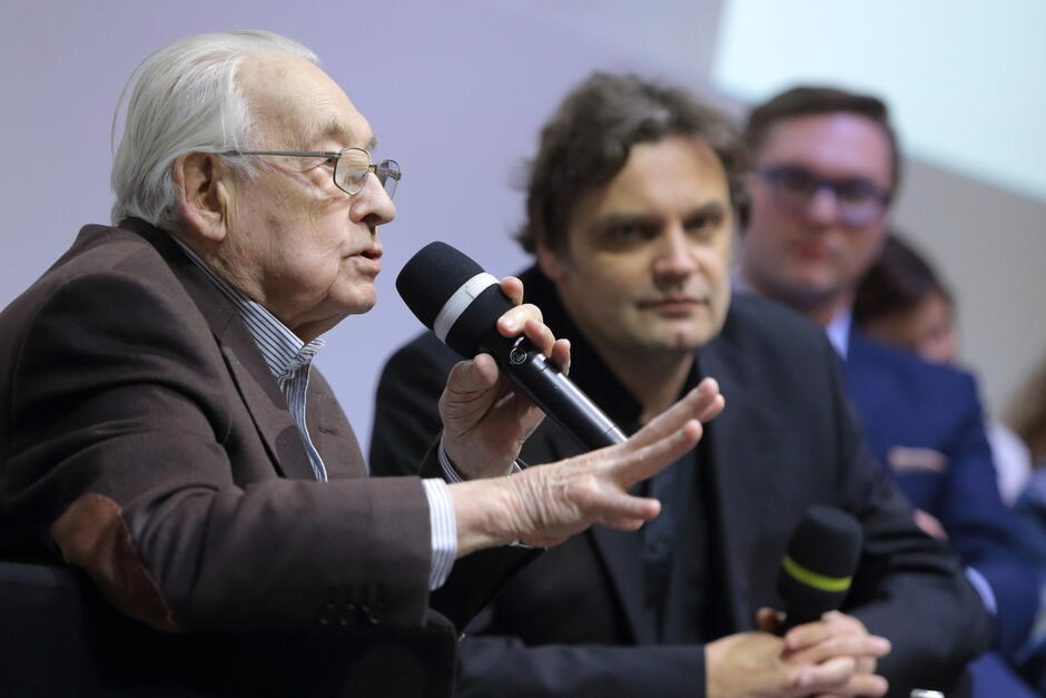 starszy człowiek, reżyser Andrzej Wajda, mówi do mikrofonu na spotkaniu, w tle ekran 