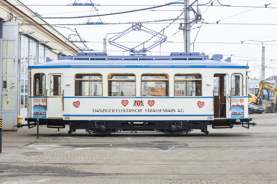 Na zdjęciu widoczny jest zabytkowy tramwaj należący do Danziger Elektrische Strassenbahn A.G., ozdobiony eleganckimi detalami i utrzymany w jasnej kolorystyce z niebieskimi akcentami. Tramwaj znajduje się na terenie zajezdni, otoczony infrastrukturą trakcyjną oraz pojazdami technicznymi.