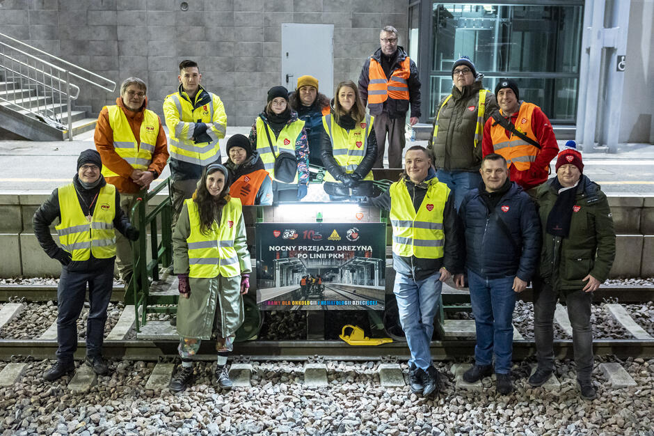 Na zdjęciu widzimy grupę osób ubranych w odblaskowe kamizelki i ciepłe ubrania, zgromadzonych wokół drezyny na torach kolejowych. Baner informuje o nocnym przejeździe drezyną po linii PKM, organizowanym w ramach akcji WOŚP na rzecz onkologii i hematologii dziecięcej.
