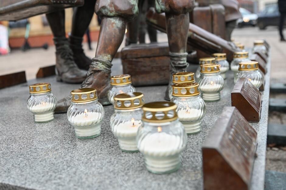 Fotografia daje zbliżenie na nogi postaci z pomnika Kindertransportu i zapalone na cokole pomnika znicze