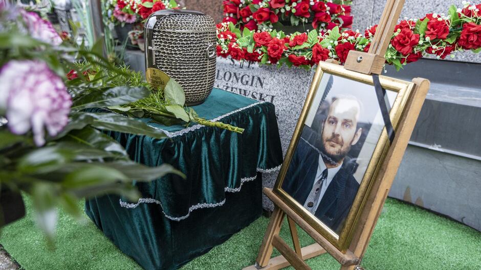 Na zdjęciu przedstawiono miejsce upamiętniające osobę zmarłą, prawdopodobnie podczas ceremonii pogrzebowej. Centralnym elementem jest urna pogrzebowa umieszczona na stoliku pokrytym zielonym, aksamitnym materiałem ozdobionym koronkowym wykończeniem. Obok urny znajduje się zdjęcie zmarłego w eleganckiej złotej ramce, ustawione na drewnianej sztaludze, z czarną wstążką symbolizującą żałobę. W tle widoczne są czerwone wieńce z róż oraz inne kwiaty, które dekorują miejsce ceremonii. Na jednym z elementów w tle można dostrzec nazwisko  Longin Lewandowski , co może sugerować, że jest to nagrobek lub tablica pamiątkowa osoby spoczywającej w tym miejscu. Całość kompozycji wskazuje na moment pożegnania i oddania czci zmarłemu.