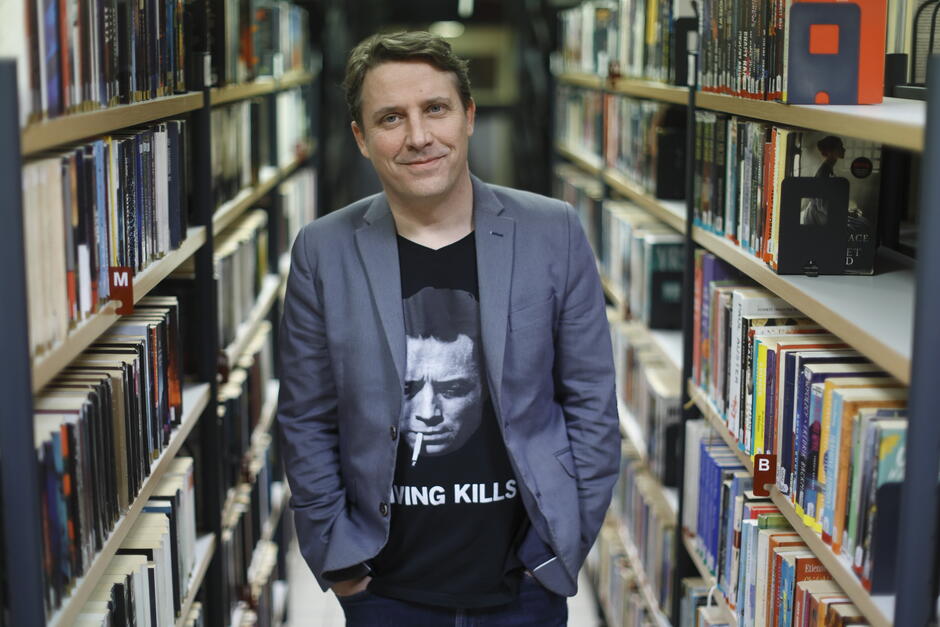 Na zdjęciu widzimy mężczyznę stojącego w alejce między regałami pełnymi książek w bibliotece. Jest ubrany w szarą marynarkę i czarną koszulkę z grafiką oraz napisem SMOKING KILLS, a na jego twarzy widnieje lekki uśmiech.