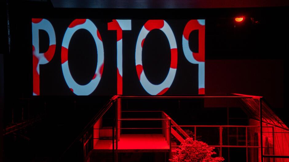 Zdjęcie przedstawia scenografię teatralną w nastrojowym czerwonym oświetleniu, na której znajduje się podwyższona konstrukcja z metalowych ram. W tle widoczny jest ekran z wyświetlonym słowem POTOP w biało-czerwonym wzorze, nawiązującym do polskiej flagi