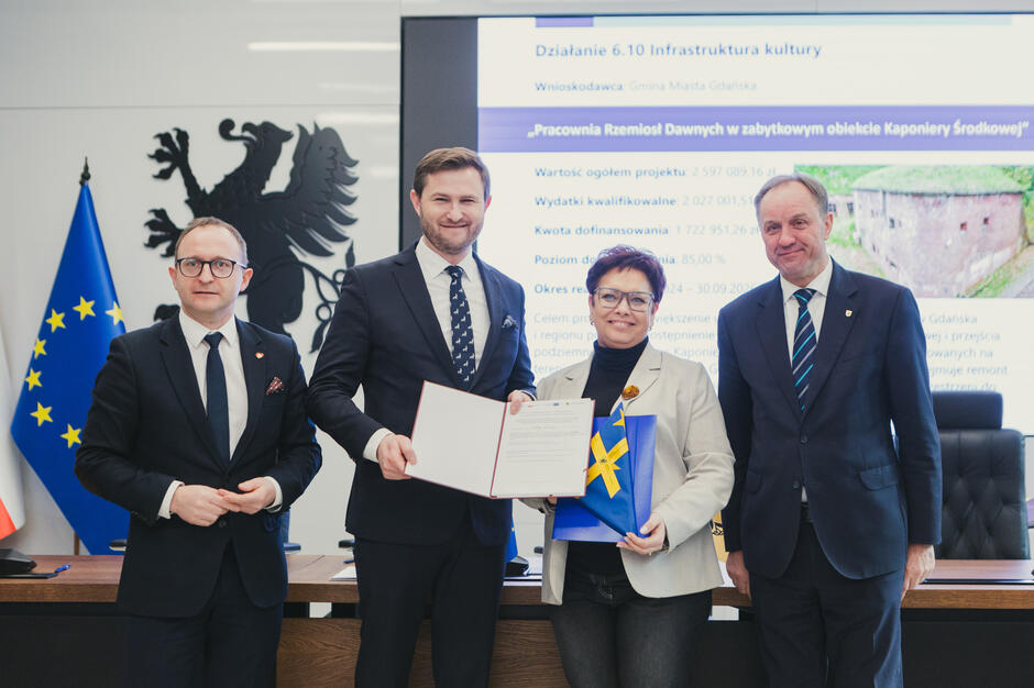 Na zdjęciu widzimy cztery osoby pozujące z dokumentem i materiałami promocyjnymi na tle flagi Unii Europejskiej oraz planszy informacyjnej dotyczącej projektu „Pracownia Rzemiosł Dawnych w zabytkowym obiekcie Kaponiery Środkowej”. Spotkanie odbywa się w oficjalnej atmosferze, a uśmiechnięci uczestnicy symbolicznie podkreślają sukces w realizacji projektu dofinansowanego z funduszy unijnych