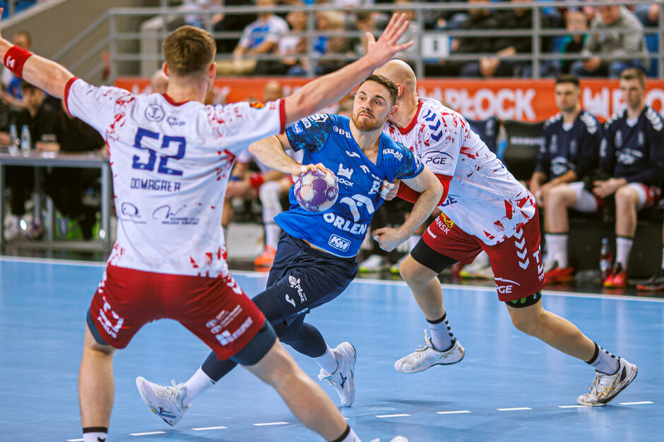 Dwóch piłkarzy ręcznych próbuje zatrzymać jednego w niebieskim stroju z piłką