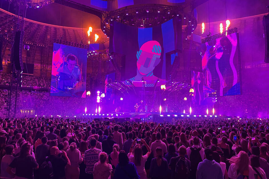 To zdjęcie przedstawia koncert w dużej, zadaszonej arenie w Gdańsku, gdzie tłum fanów ogląda widowiskowy występ artysty na imponującej scenie z efektami świetlnymi, ogniem i animacjami na ekranach. Dominują fioletowe i niebieskie światła, a na głównym ekranie wyświetlana jest futurystyczna grafika oraz zbliżenia na muzyków