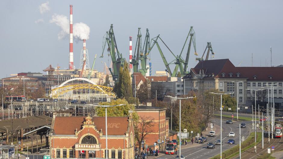 To zdjęcie przedstawia widok na Stocznię Gdańską i jej otoczenie. Na pierwszym planie widoczny jest budynek z czerwonej cegły, na którym znajduje się logo KFC. W tle znajdują się charakterystyczne zielone dźwigi stoczniowe, a także kominy emitujące biały dym. Po lewej stronie można zauważyć napis „STOCZNIA GDAŃSKA” na jednym z budynków przemysłowych. Widoczne są także ulice z samochodami, tramwajami i pieszymi, a nad torami kolejowymi widać żółty łukowy most. Całość ukazuje industrialny krajobraz Gdańska, będący połączeniem historii, przemysłu i nowoczesnej infrastruktury miejskiej.