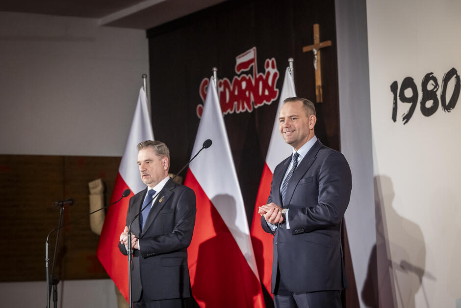 Dwaj mężczyźni w garniturach na scenie, w tle biało-czerwone flagi