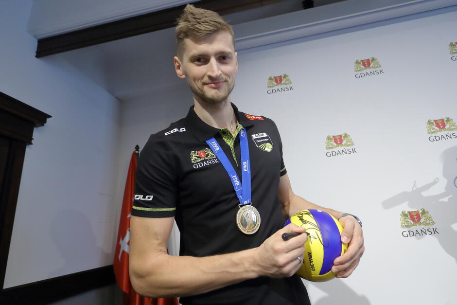 Mężczyzna w koszulce sportowej z medalem na szyi i piłką w rękach, na której składa autograf