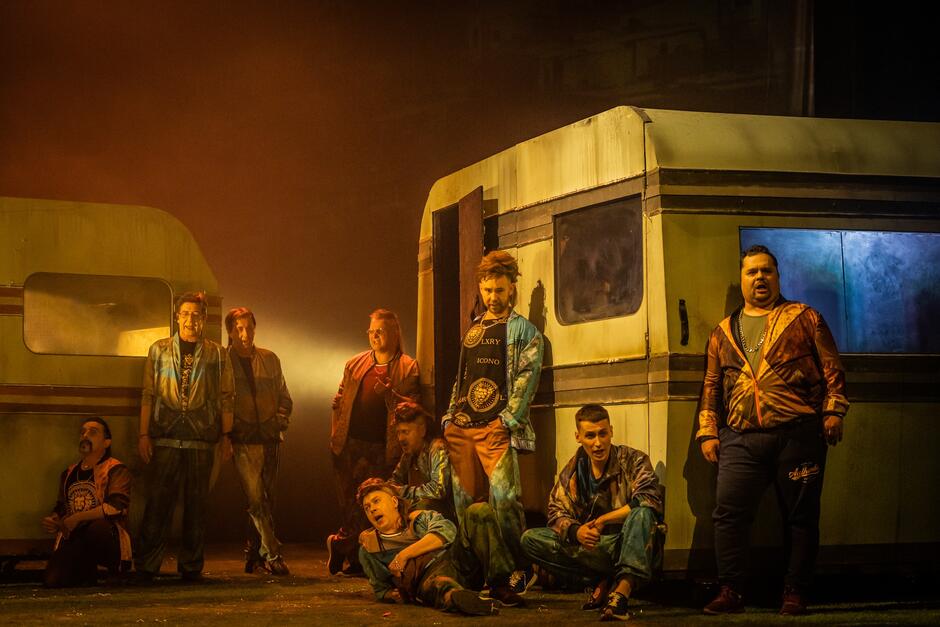 Zdjęcie przedstawia grupę mężczyzn w barwnych, ulicznych strojach stojących i siedzących wokół starych przyczep kempingowych w klimatycznym, teatralnym oświetleniu