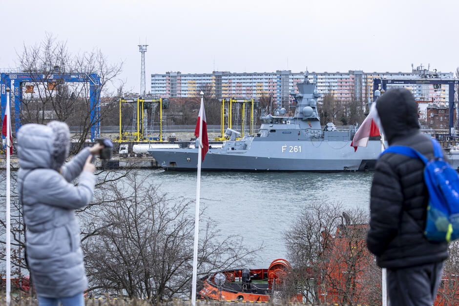 Okręt na rzece, przed nimi dwie osoby, które z nabrzeża robią zdjęcia