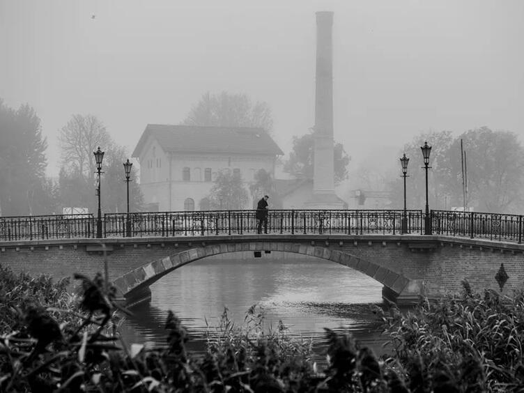 Zdjęcie przedstawia malowniczy most z kutymi barierkami i latarniami, rozciągający się nad spokojną rzeką, na którym stoi samotna postać w ciemnym płaszczu. W tle, spowity gęstą mgłą, widać budynek z dwuspadowym dachem oraz wysoką, wąską wieżę przypominającą komin fabryczny, co nadaje scenie nostalgiczny i tajemniczy nastrój