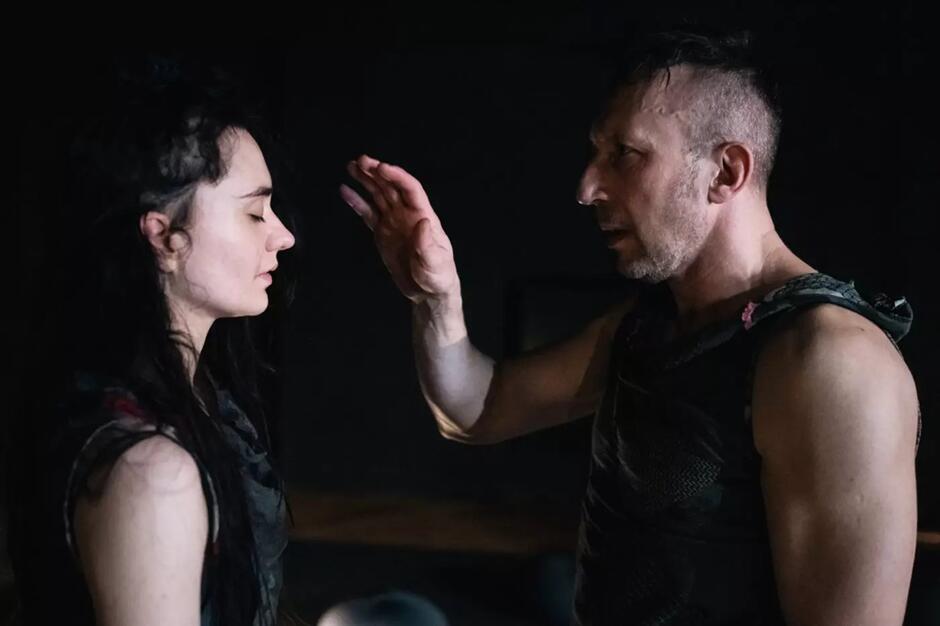 Na zdjęciu widzimy scenę teatralną lub artystyczną, w której mężczyzna z wygoloną bokiem głową, ubrany w postapokaliptyczny strój, unosi rękę w kierunku kobiety o długich, ciemnych włosach, mającej zamknięte oczy