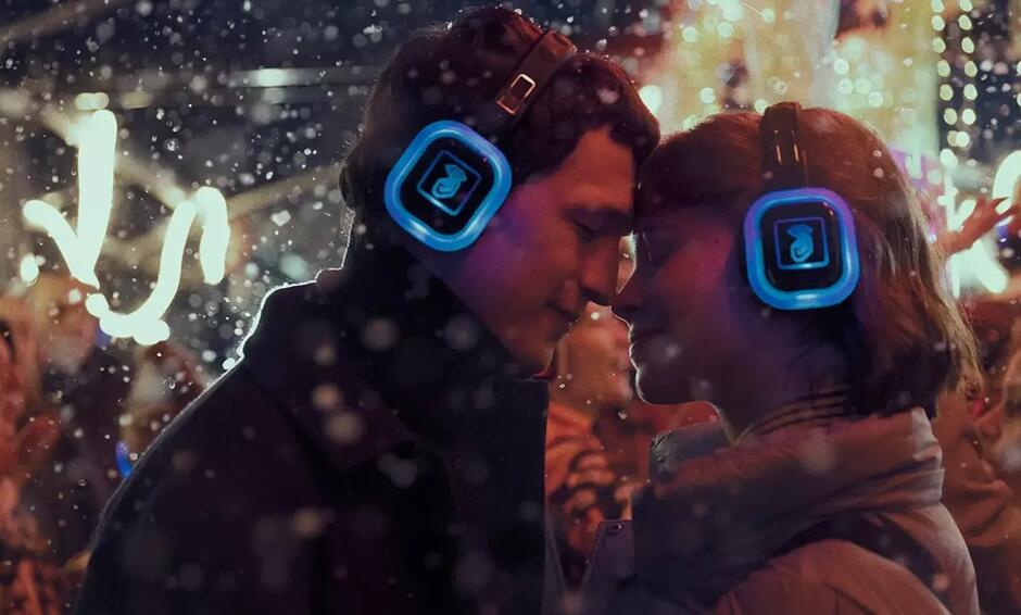 Na zdjęciu widzimy zakochaną parę, która stoi blisko siebie w zimowej scenerii, otoczona padającym śniegiem i świątecznymi światłami. Oboje noszą świecące na niebiesko słuchawki, sugerujące udział w cichej imprezie (silent disco), co tworzy intymną i magiczną atmosferę