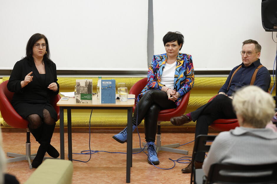Na zdjęciu widać spotkanie autorskie, w którym uczestniczą trzy osoby siedzące na czerwonych fotelach, a jedna z nich gestykuluje podczas wypowiedzi. Przed nimi stoi stolik z książkami, szklanką wody i notatkami