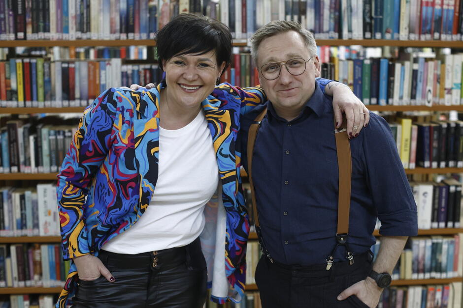 Na zdjęciu widać uśmiechniętych kobietę i mężczyznę pozujących razem na tle bibliotecznych regałów pełnych książek. Kobieta w kolorowej marynarce obejmuje mężczyznę, który ma na sobie granatową koszulę i szelki