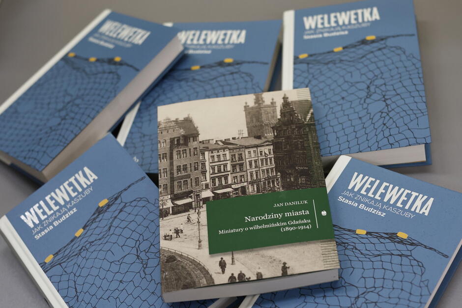 Na zdjęciu widocznych jest kilka egzemplarzy książki Welewetka. Jak znikają Kaszuby autorstwa Stasi Budzisz oraz jedna książka Narodziny miasta. Miniatury o wilhelmińskim Gdańsku (1890-1914) Jana Daniluka. Książki leżą na stole, a ich okładki przyciągają uwagę – jedna z nich przedstawia historyczną fotografię miasta, a pozostałe mają charakterystyczny niebieski design z motywem sieci rybackiej.