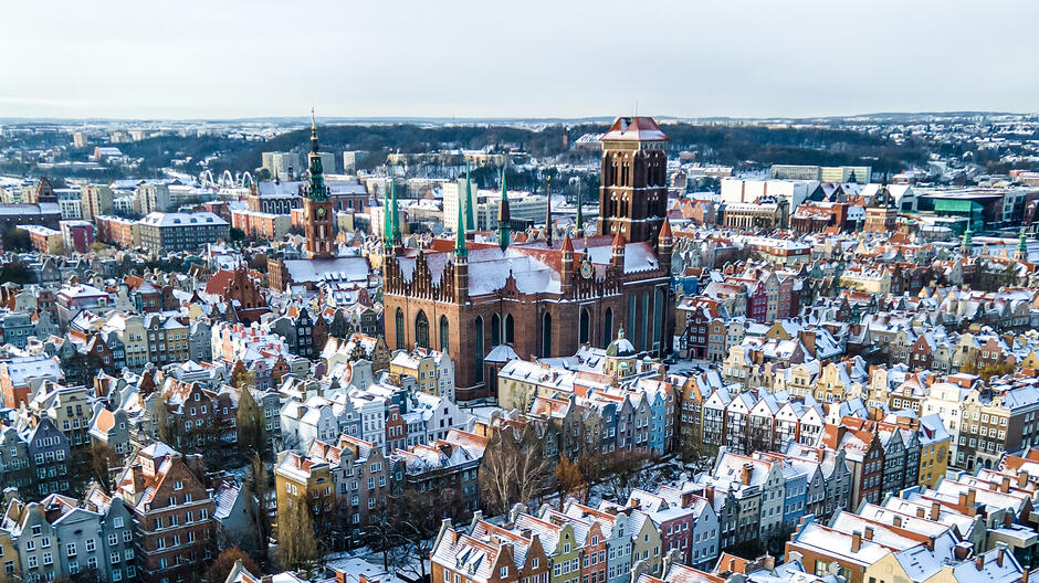 Na zdjęciu widoczna jest zimowa panorama miasta Gdańsk. Charakterystyczne budynki z cegły, w tym kościoły i wieże zegarowe, pokryte są delikatną warstwą śniegu. Wśród nich wyróżnia się bazylika Mariacka – największy kościół z cegły na świecie, z charakterystyczną masywną wieżą po prawej stronie obrazu. Po lewej stronie można dostrzec Ratusz Głównego Miasta z wysoką wieżą oraz inne zabytkowe budynki. Na pierwszym planie widać również budynek dworca kolejowego Gdańsk Główny z zielonymi wieżyczkami i zegarem. W tle, oprócz nowoczesnych budynków, znajdują się także wiatraki, wskazujące na obecność farmy wiatrowej. Niebo jest zachmurzone, a cała sceneria ma chłodny, zimowy klimat. Śnieg na dachach i drzewach dodaje uroku temu historycznemu widokowi.