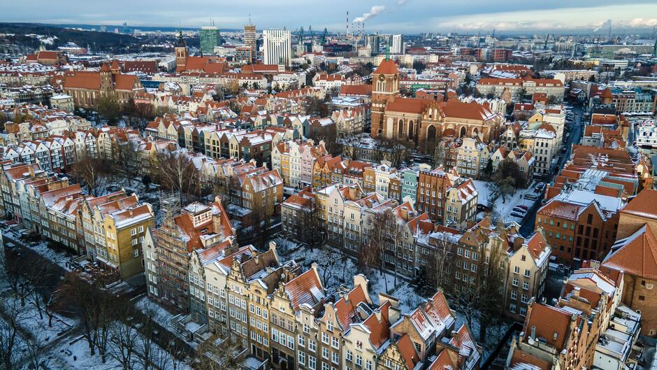 Obraz przedstawia zimowy widok na miasto z lotu ptaka. Można zauważyć urokliwe, historyczne kamienice z charakterystycznymi czerwonymi dachami pokrytymi cienką warstwą śniegu. W tle widoczne są nowoczesne budynki, industrialne obiekty oraz port z dźwigami, co wskazuje na duże miasto o bogatej historii i rozwiniętej gospodarce.