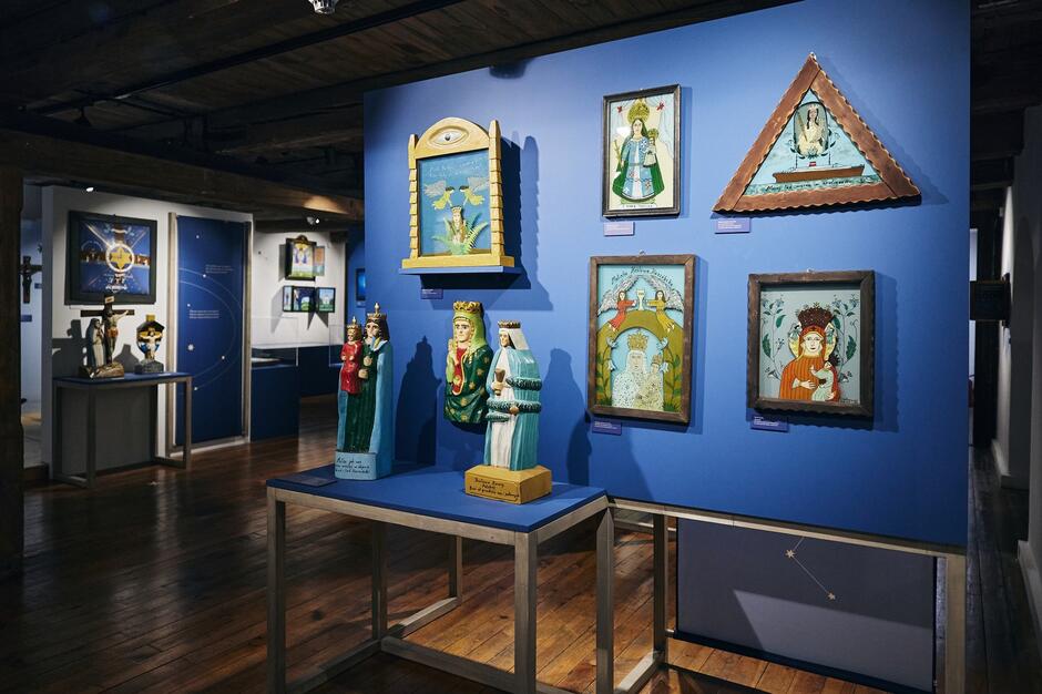 Zdjęcie przedstawia wnętrze muzeum lub galerii sztuki, w którym eksponowane są rzeźby i obrazy o ludowym, sakralnym charakterze, umieszczone na tle niebieskich ścian. Widoczne dzieła przedstawiają religijne postacie, takie jak Matka Boska i święci, wykonane w stylu naiwnej sztuki ludowej, co nadaje ekspozycji unikalny i tajemniczy klimat