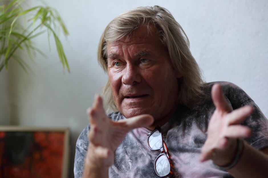 Na zdjęciu znajduje się mężczyzna o długich, blond włosach, który intensywnie gestykuluje rękami podczas rozmowy. Ma na sobie koszulkę w stylu tie-dye, a na szyi zawieszone okulary, co nadaje mu swobodny, artystyczny wygląd