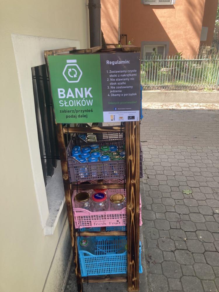 Na zdjęciu widzimy "Bank Słoików" – inicjatywę umożliwiającą wymianę słoików. Jest to drewniany regał ustawiony na zewnątrz, wyposażony w plastikowe skrzynki z różnymi słoikami, głównie z nakrętkami. Na górze regału znajduje się zielona tabliczka z logo przedstawiającym słoik i napisem „BANK SŁOIKÓW – zabierz/przynieś, podaj dalej”. Obok widnieje regulamin, który zawiera zasady korzystania z inicjatywy, m.in. konieczność zostawiania czystych słoików z nakrętkami, dbanie o porządek oraz zakaz zostawiania jedzenia.