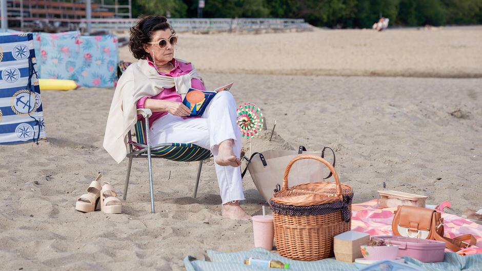 Na zdjęciu widzimy elegancką starszą kobietę siedzącą na plaży w rozkładanym krześle, czytającą książkę. Otacza ją piknikowy ekwipunek, w tym kosz wiklinowy, torby, jedzenie oraz kolorowy koc