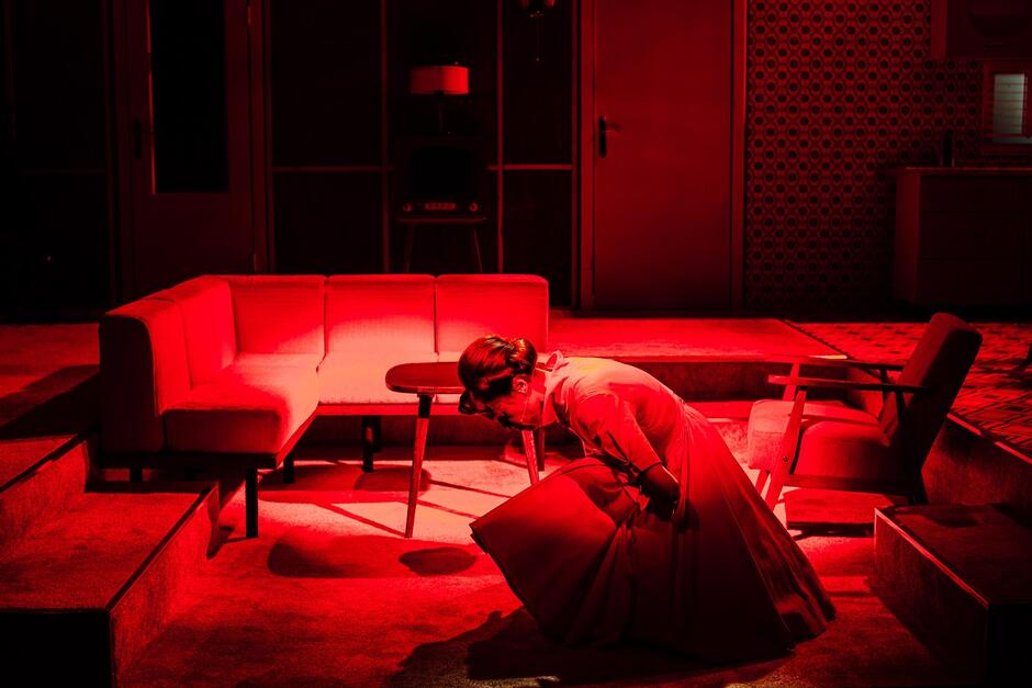Scena teatralna oświetlona na czerwono, pośrodku kuca zgięta w pół kobieta w sukience a la lata 60