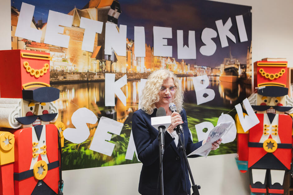 Na zdjęciu widać kobietę w eleganckim stroju, stojącą przy mikrofonie i przemawiającą do publiczności podczas wydarzenia. Za nią znajduje się dekoracja z dużymi literami układającymi się w napis „LETNIEWSKI KLUB SENIORÓW” na tle nocnej panoramy Gdańska, a po bokach stoją duże figurki dziadków do orzechów