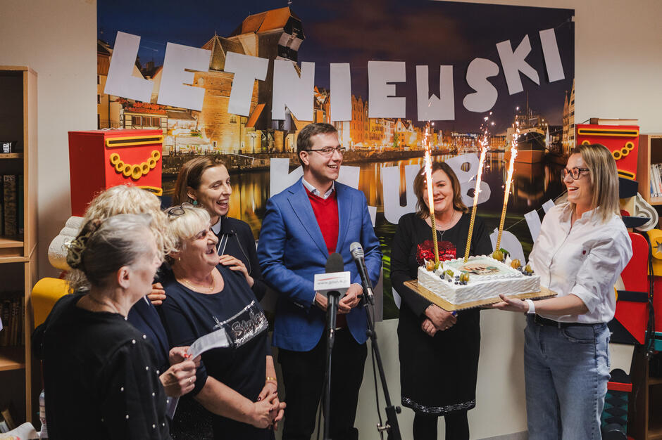 Na zdjęciu grupa uśmiechniętych ludzi świętuje w pomieszczeniu udekorowanym napisem „LETNIEWSKI KLUB SENIORA”, a jedna z osób trzyma tort z płonącymi fontannami. W centrum uwagi jest mężczyzna w niebieskiej marynarce i czerwonym swetrze, otoczony przez roześmiane kobiety, co sugeruje radosną atmosferę wydarzenia