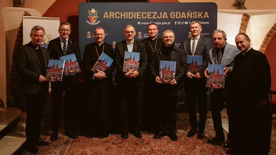 Na zdjęciu widzimy grupę mężczyzn pozujących do wspólnej fotografii podczas wydarzenia związanego z Archidiecezją Gdańską. Stoją oni na tle ścianki z nazwą i herbem archidiecezji oraz informacjami o jej stronie internetowej i mediach społecznościowych. Osoby na zdjęciu to zarówno duchowni, jak i świeccy, a każdy z nich trzyma w rękach egzemplarz książki z okładką przedstawiającą kościół. Uroczysta atmosfera, eleganckie stroje oraz historyczny wystrój wnętrza sugerują, że wydarzenie odbywa się w kościelnym lub zabytkowym budynku.  