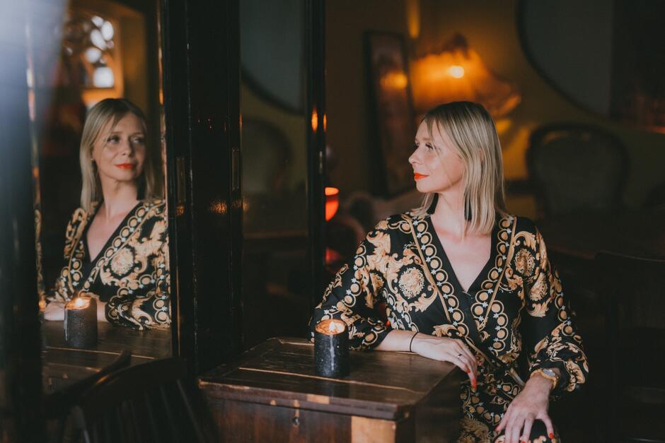 Na zdjęciu znajduje się elegancko ubrana kobieta siedząca w przytulnym, klimatycznym wnętrzu, oświetlonym ciepłym światłem lamp i świecy. Jej odbicie w lustrze tworzy ciekawy efekt wizualny, dodając głębi i tajemniczości do kompozycji