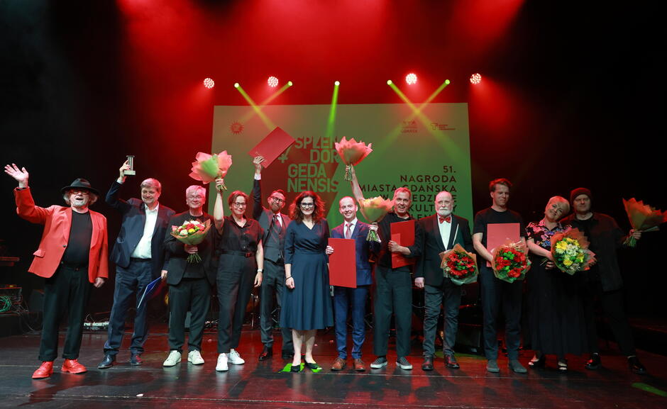 Na zdjęciu widać grupę osób stojących na scenie podczas gali Splendor Gedanensis 2024, prestiżowej nagrody miasta Gdańska w dziedzinie kultury. Uczestnicy, elegancko ubrani, trzymają dyplomy, statuetki i bukiety kwiatów, a scena oświetlona jest efektownym czerwonym światłem, tworząc uroczystą atmosferę.