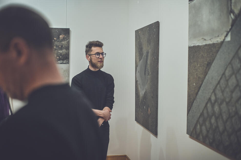 Zdjęcie przedstawia scenę z wernisażu lub wystawy sztuki, gdzie młody mężczyzna w czarnym golfie i okularach ogląda abstrakcyjne obrazy przedstawiające tekstury betonu i asfaltu. W tle widać innych uczestników wydarzenia, co nadaje zdjęciu atmosferę skupienia i artystycznej refleksji