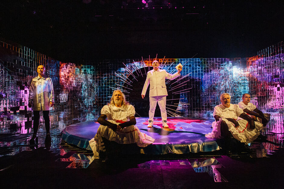 Zdjęcie przedstawia scenę teatralną o futurystycznym, surrealistycznym charakterze, z lustrzaną, wielobarwną scenografią odbijającą światła. Na środku dominuje postać ubrana na biało, stojąca na podwyższeniu w otoczeniu aktorów w kontrastujących, stylizowanych kostiumach, co sugeruje ekspresyjne, symboliczne przedstawienie